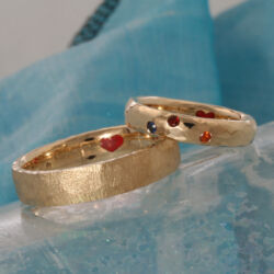 Een set relatieringen. ieder een eigen stijl, maar toch een set. Hij ruw en mat, zij hamerslag glanzend en drie edelstenen: saffier, robijn en hessoniet. In de ringen allebei een rood hartje. Ruw en hamerslag