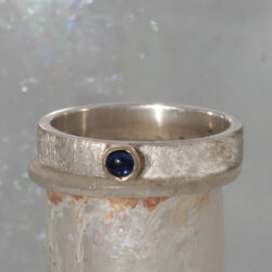 Een ijsmatte zilveren ring met een ronde saffier, gezet in een geelgouden zetkastje. De ring kan als aanschuifring gecombineerd worden. Prijs: €345,-